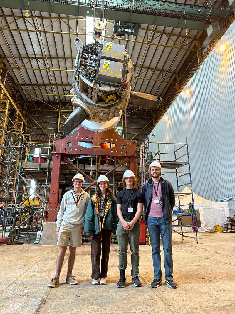 Four students wearing hard hats standing in front of a tidal turbine