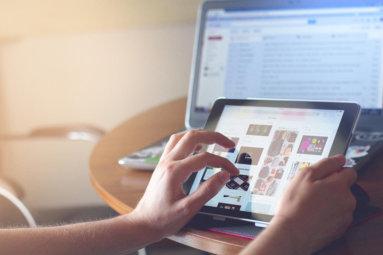 Hands holding an iPad with a laptop on a table in the background