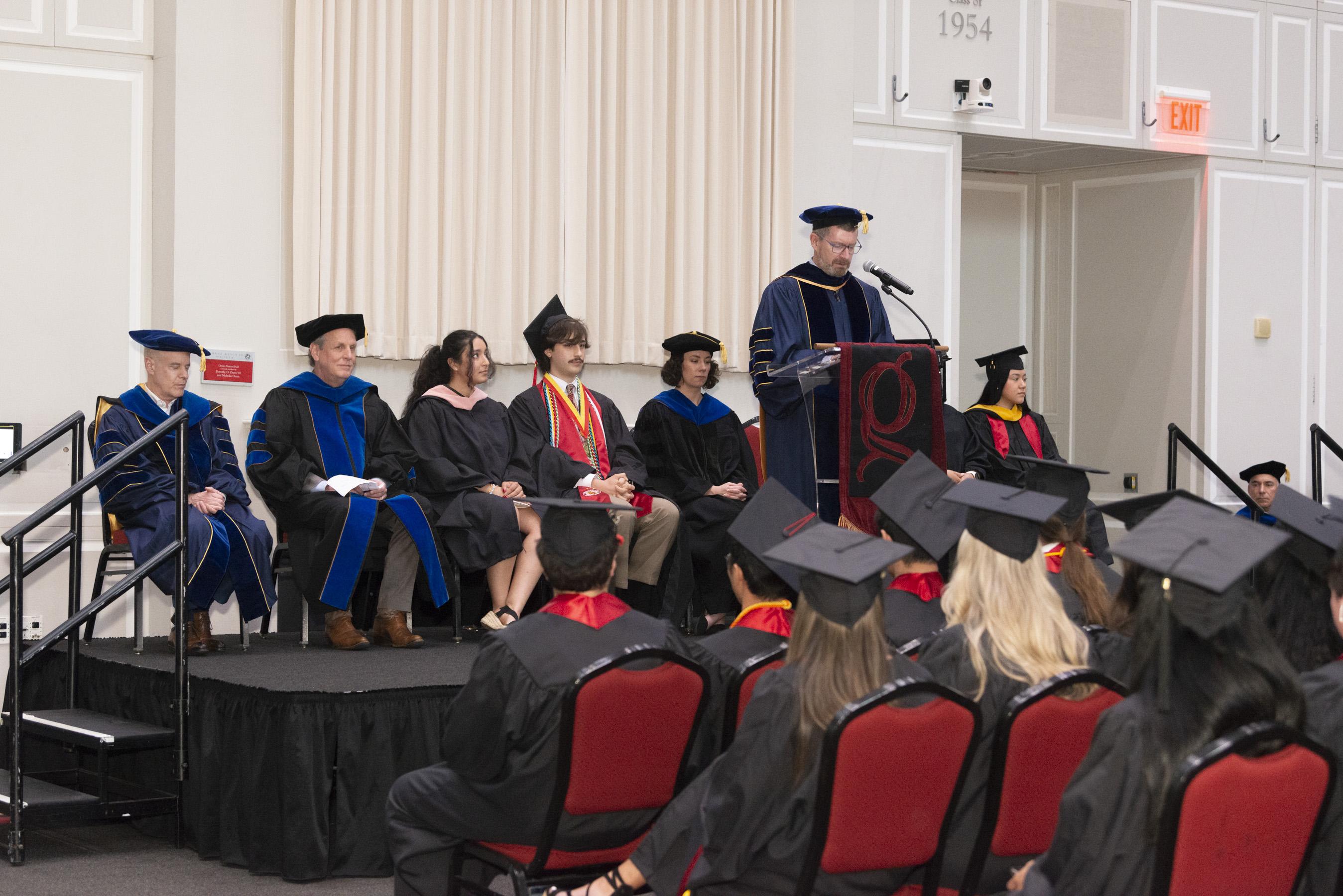 Gemstone staff and faculty wearing regalia sitting on a stage with a podium