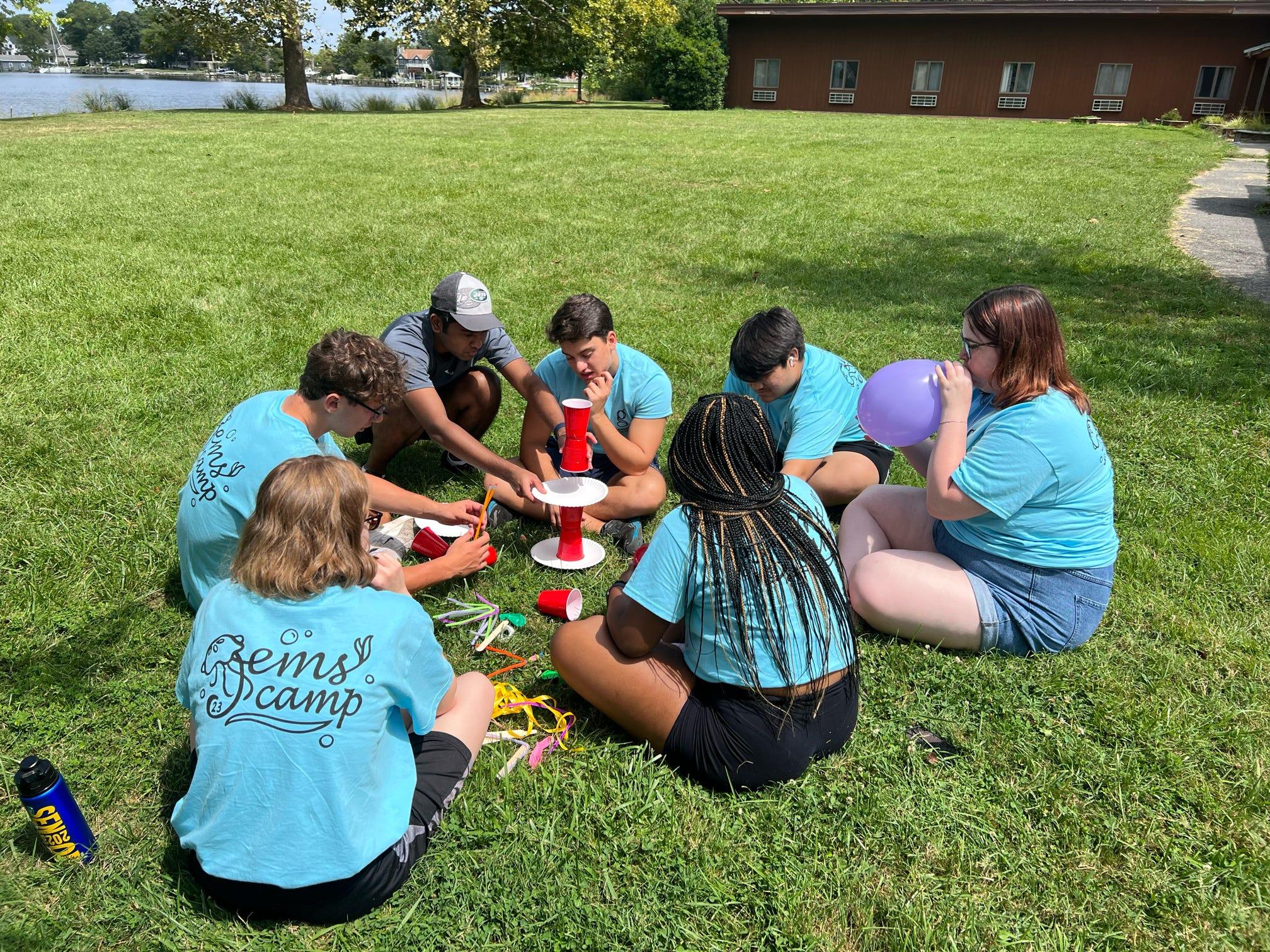 Students sitting on grass building tower of paper plates and plastic cups