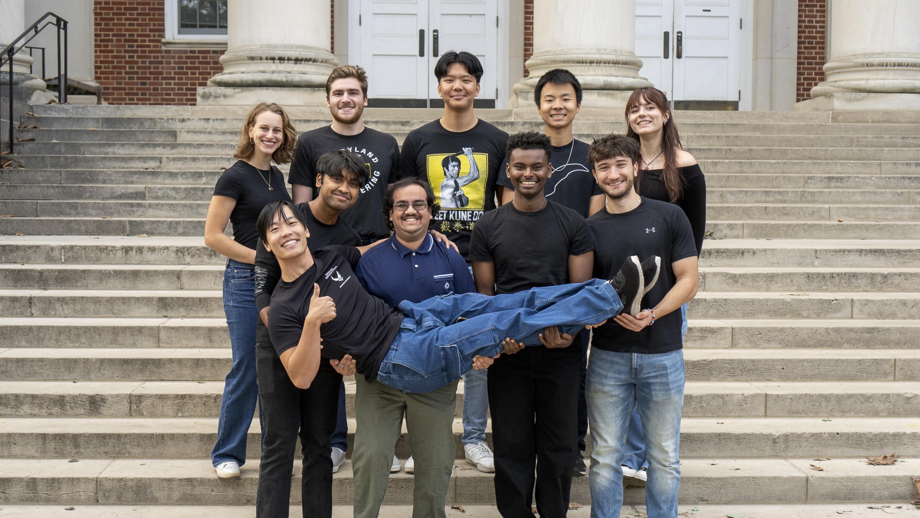 Group of students posing in front of stairs and picking one team member up