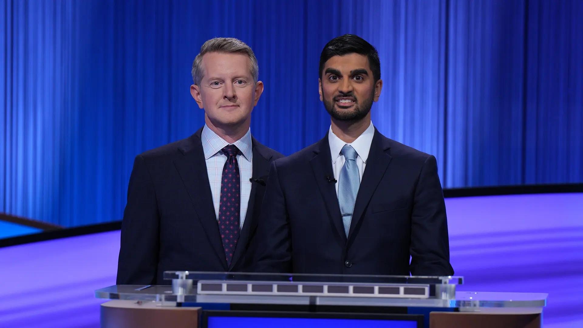 Two men in suits standing in front of blue curtain at Jeopardy game show