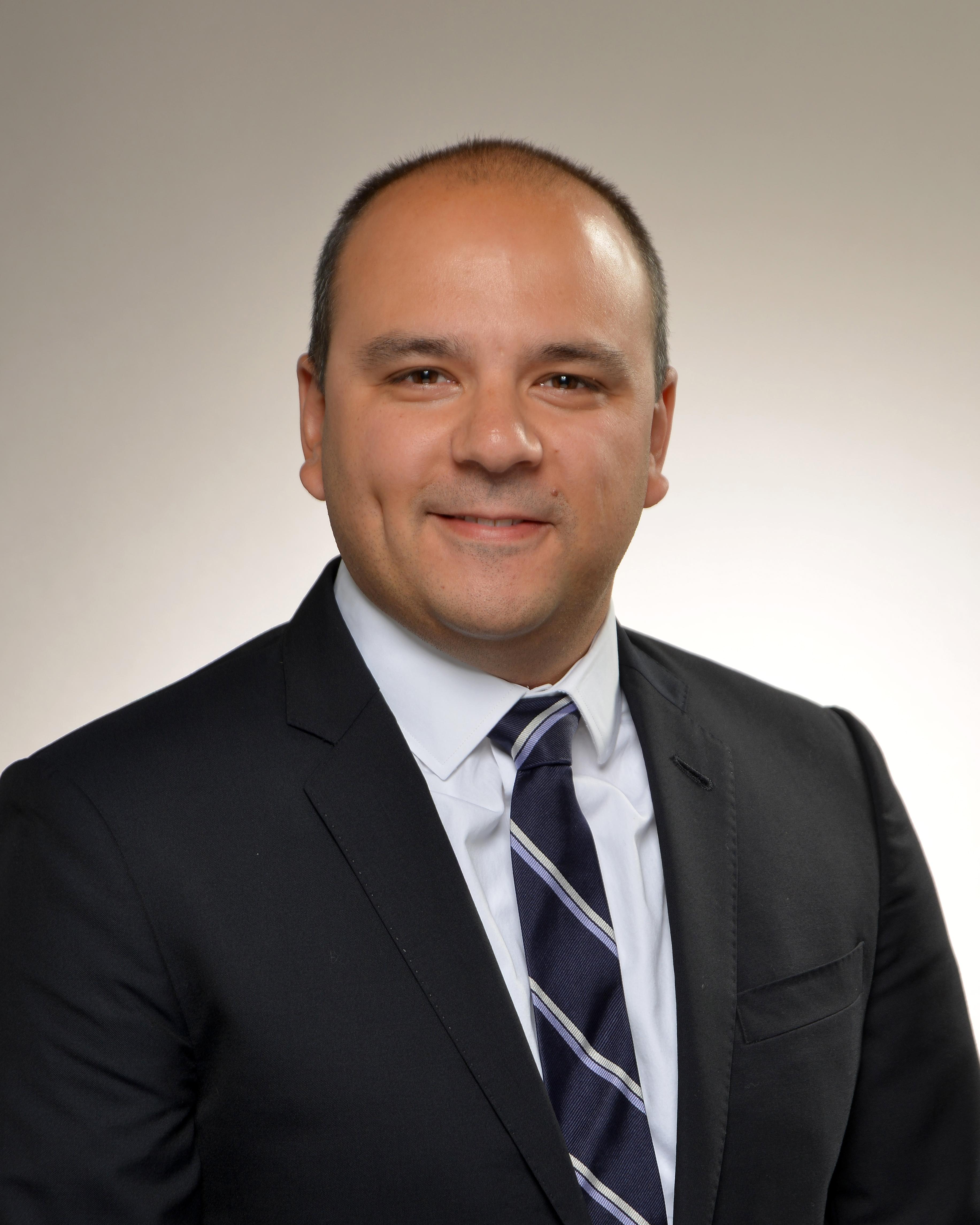 Photo of Greg Teitelbaum in black suit, white shirt, and navy and white tie against a gradient grey background