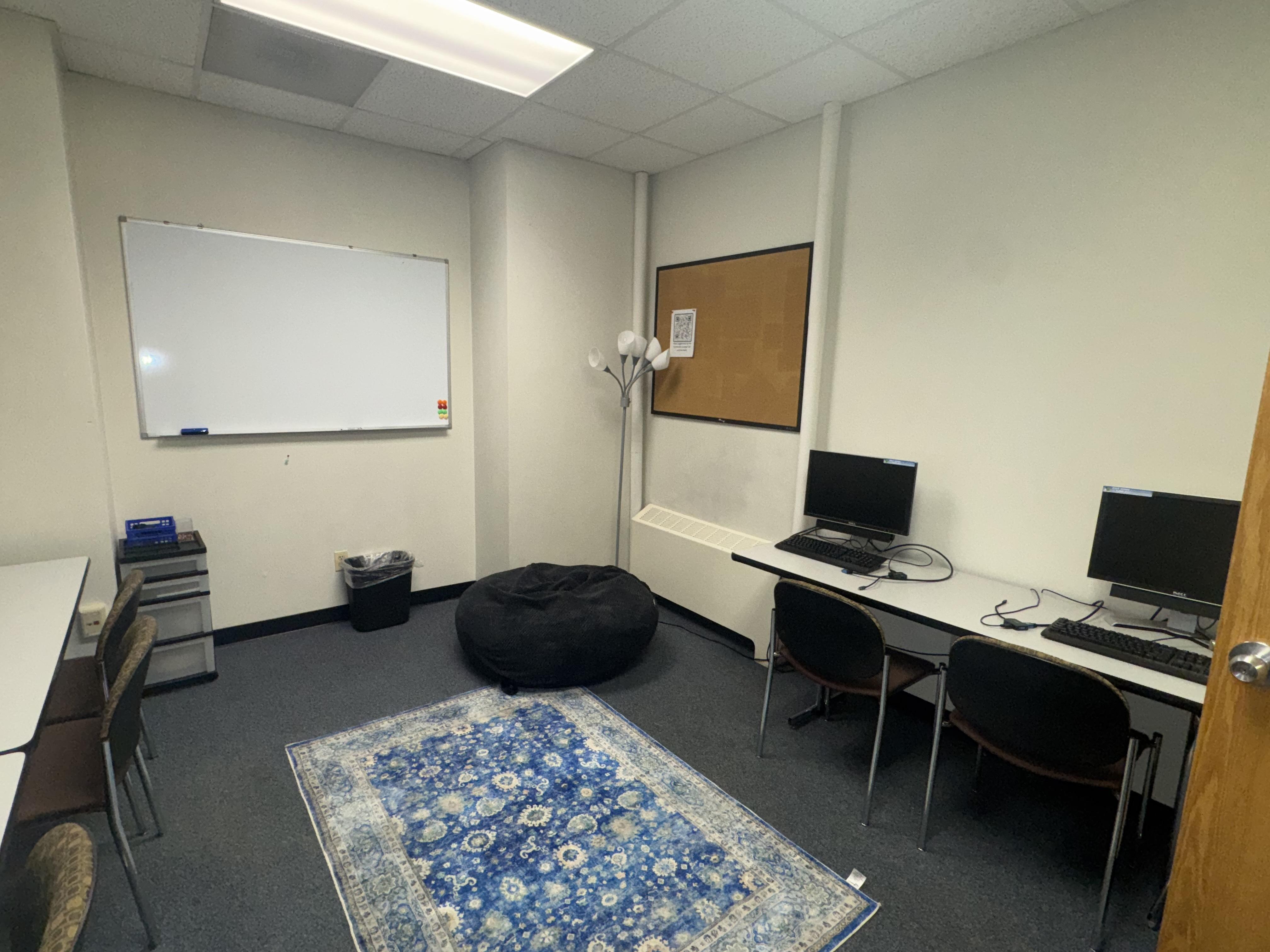 Photo of right side of commuter lounge that shows two more tables along the wall, chairs, monitors, and keyboards
