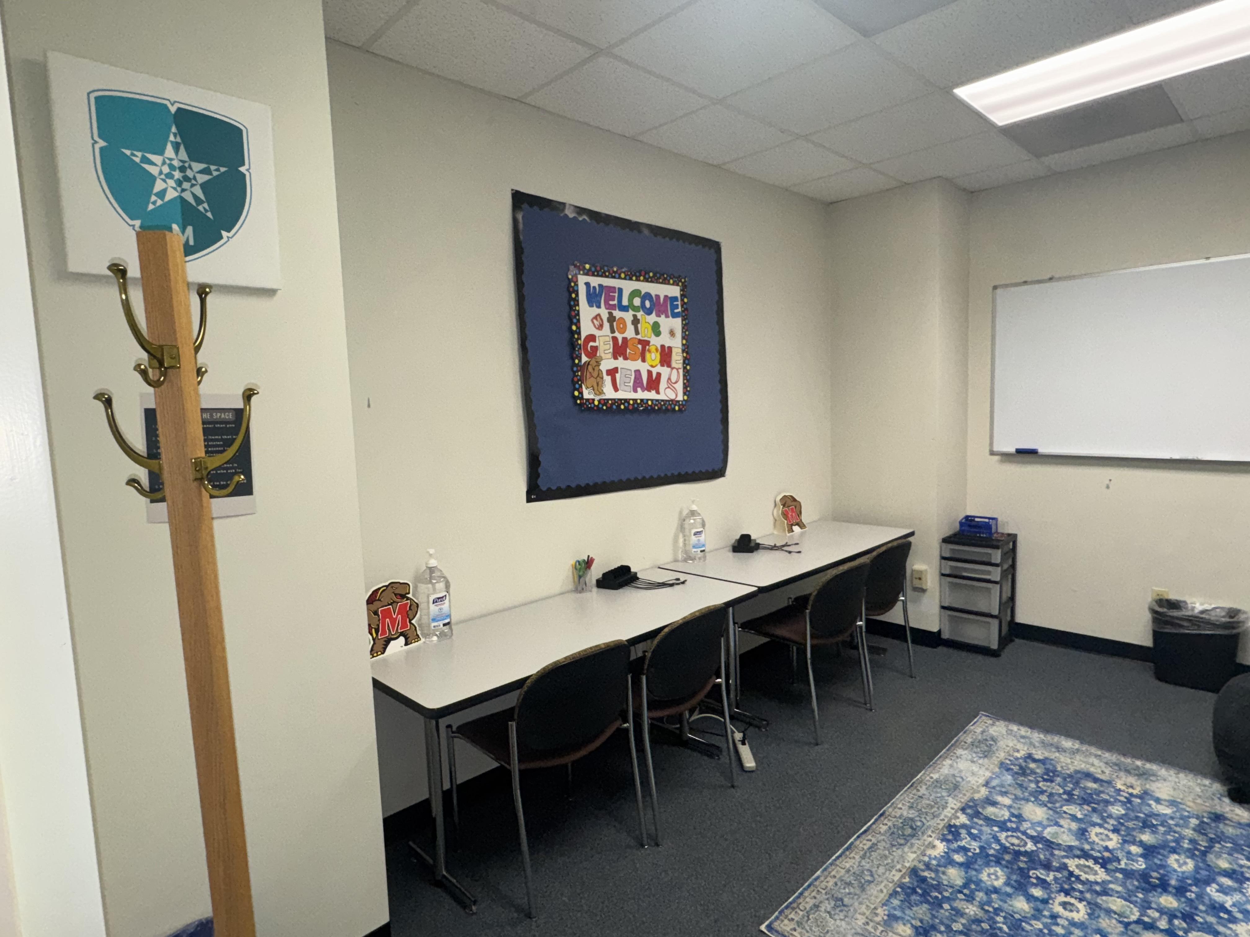 Photo of left side of commuter lounge that shows coat rack, bulletin board, two long tables along the wall, chairs, whiteboard, and a rug on the floor
