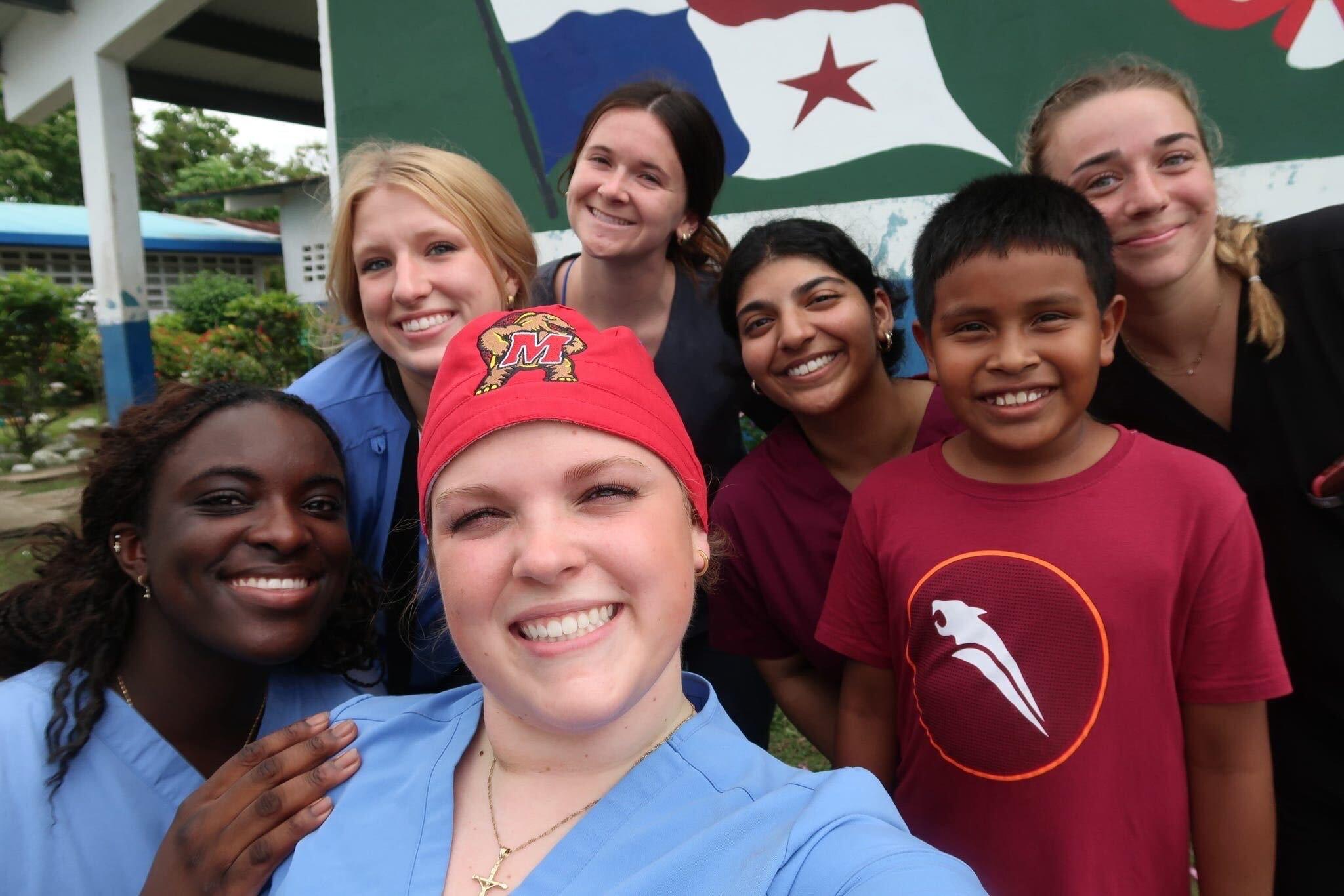 Three college students wearing medical scrubs posing and smiling with group of kids